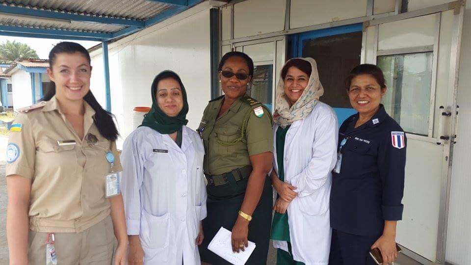 A group of diverse female professionals, including military officers and medical doctors in lab coats, standing together outside a medical facility representing international cooperation.